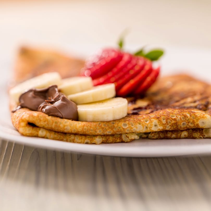 European crepe topped with Nutella, sliced bananas, and strawberries on a white plate.