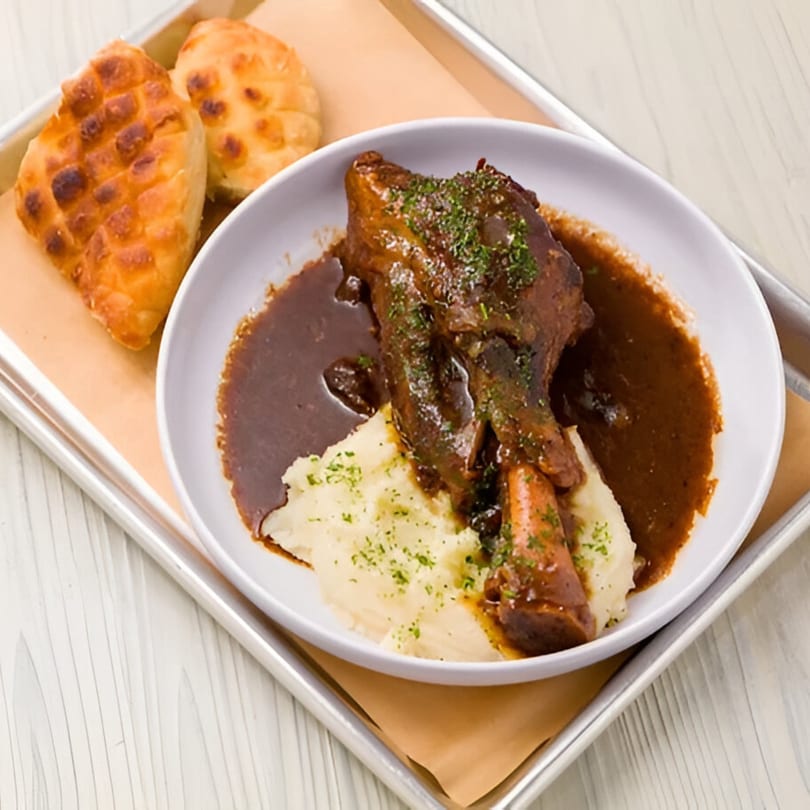 Braised lamb shank with mashed potatoes and gravy, served with two pieces of bread.