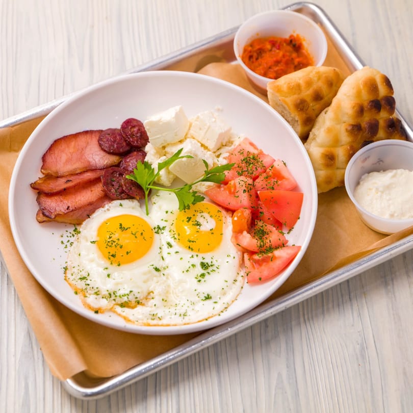 A Cafe Balkan breakfast platter with fried eggs, bacon, sausage, feta, tomatoes, bread, and dips.